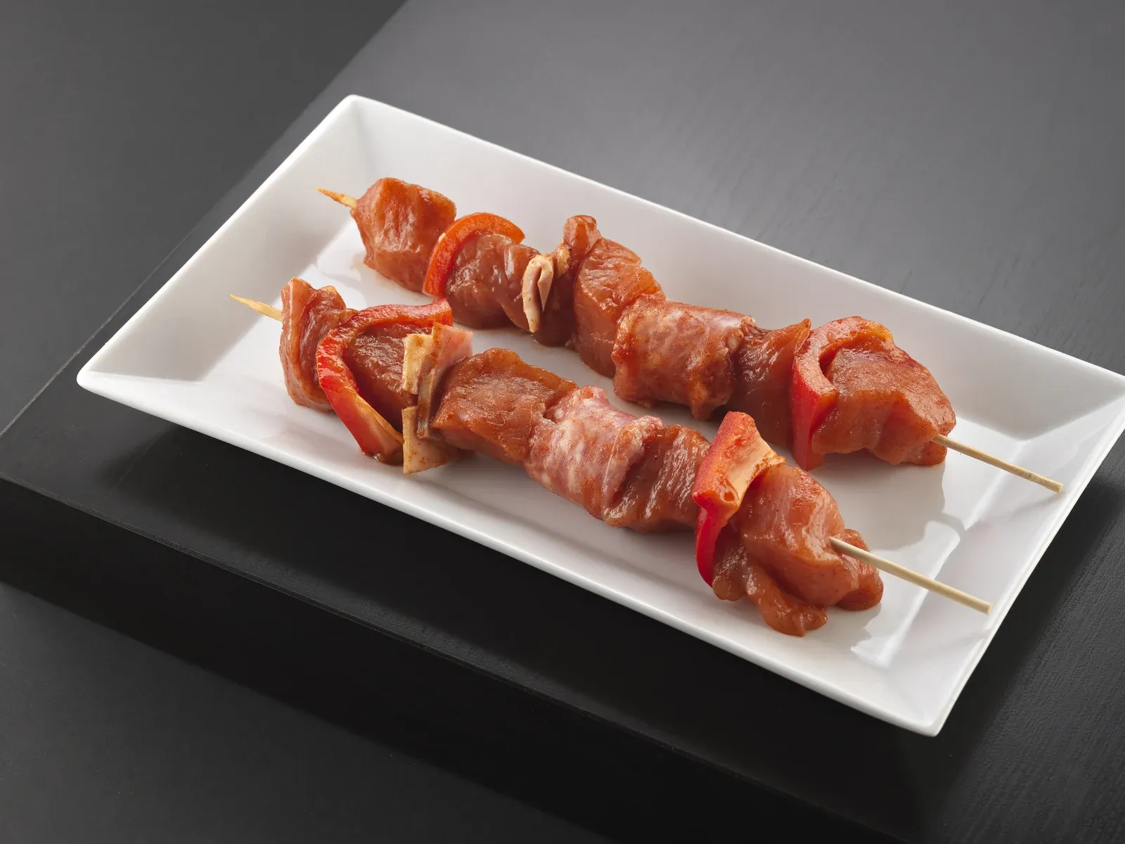Dues broquetes crues de carn adobada amb pebrot vermell, en una safata blanca